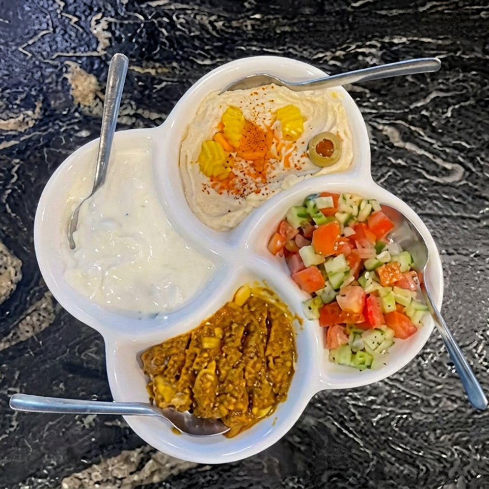 Compartmentalized platter with hummus, creamy dip, chopped salad, and spiced lentils on a black marble table.