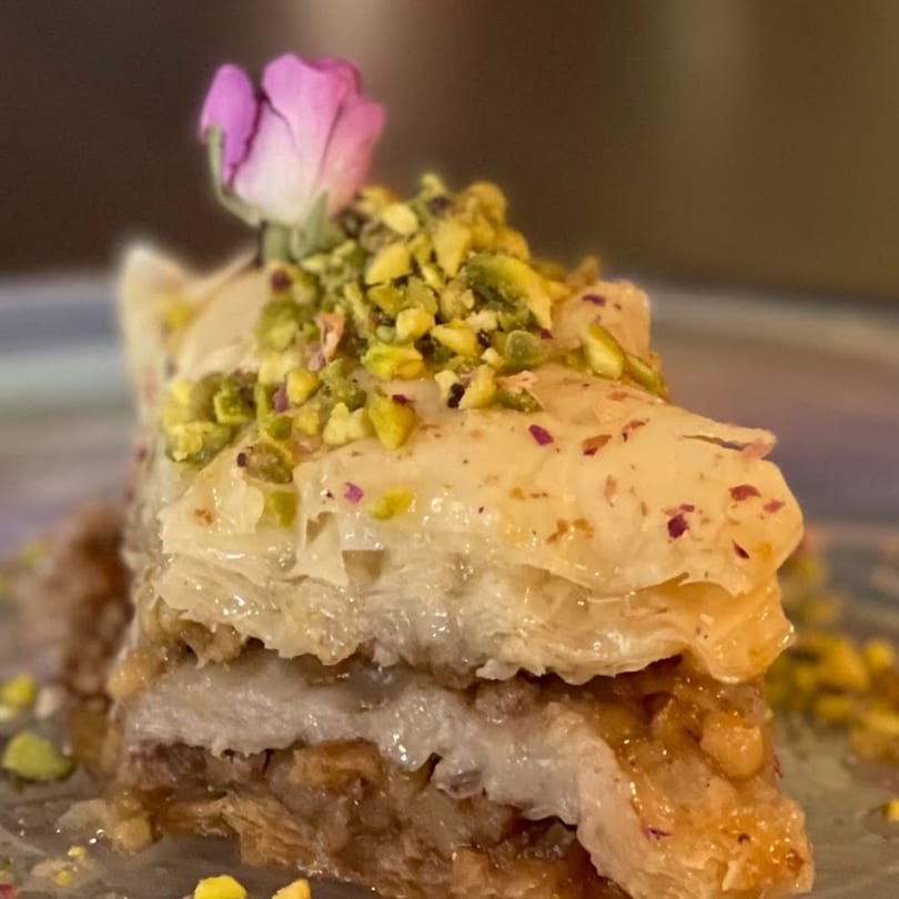 Close-up of a slice of Persian baklava, garnished with pistachios and a pink rosebud.