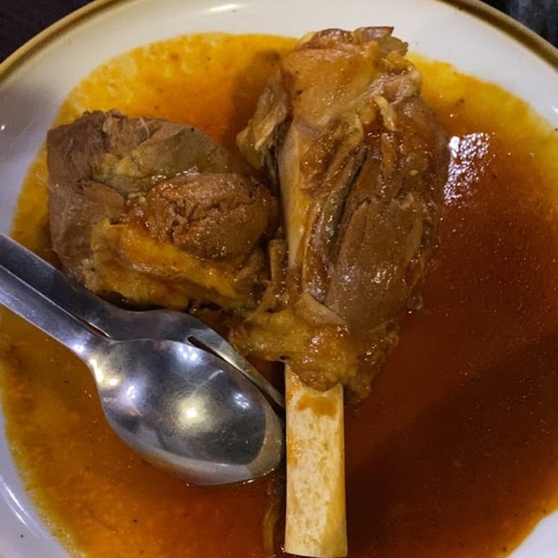 Two succulent lamb shanks in a rich, savory broth, served with a spoon and fork.